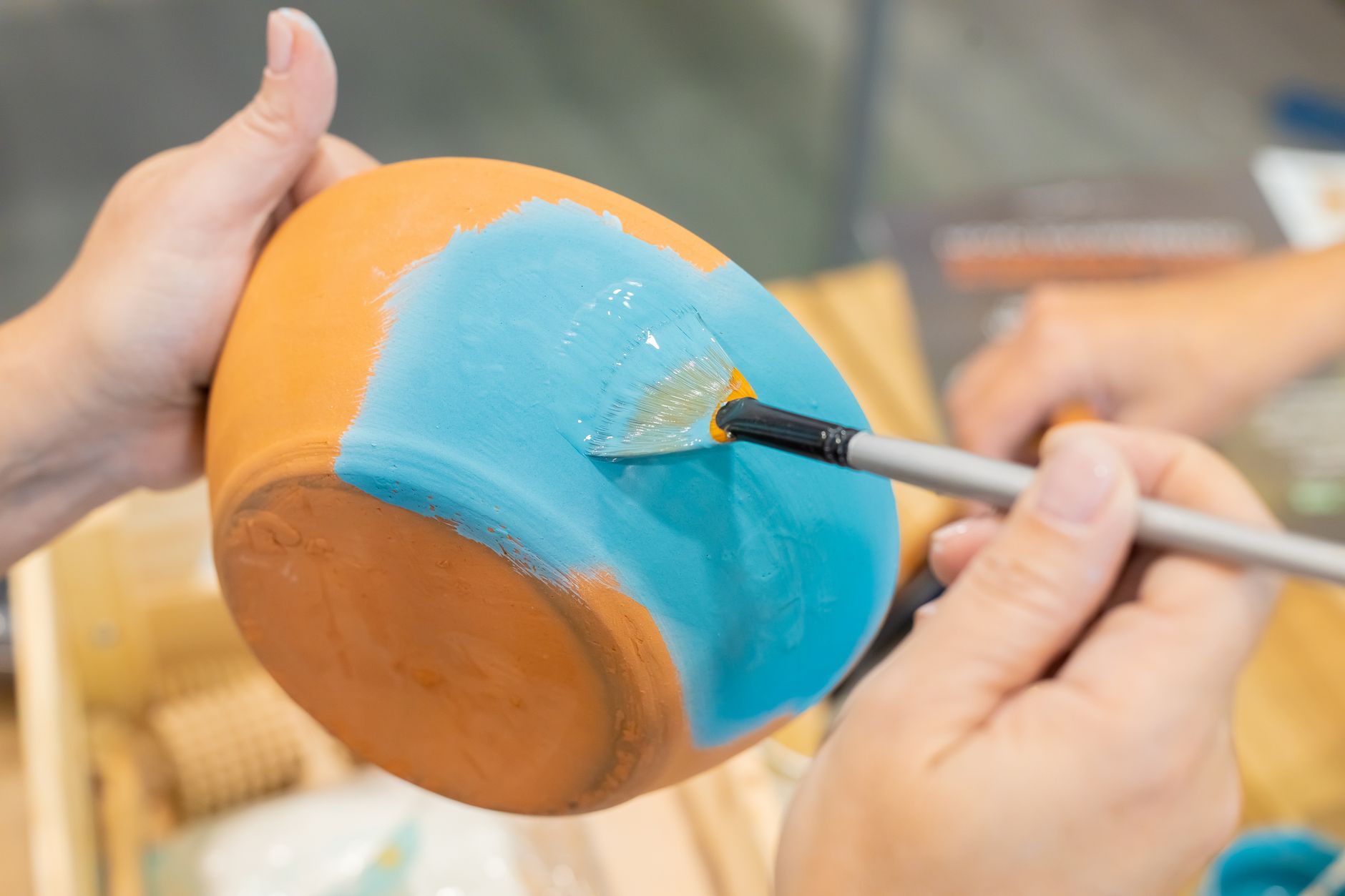 Mains peignant en bleu turquoise un pot en terre cuite retourné avec un pinceau éventail.