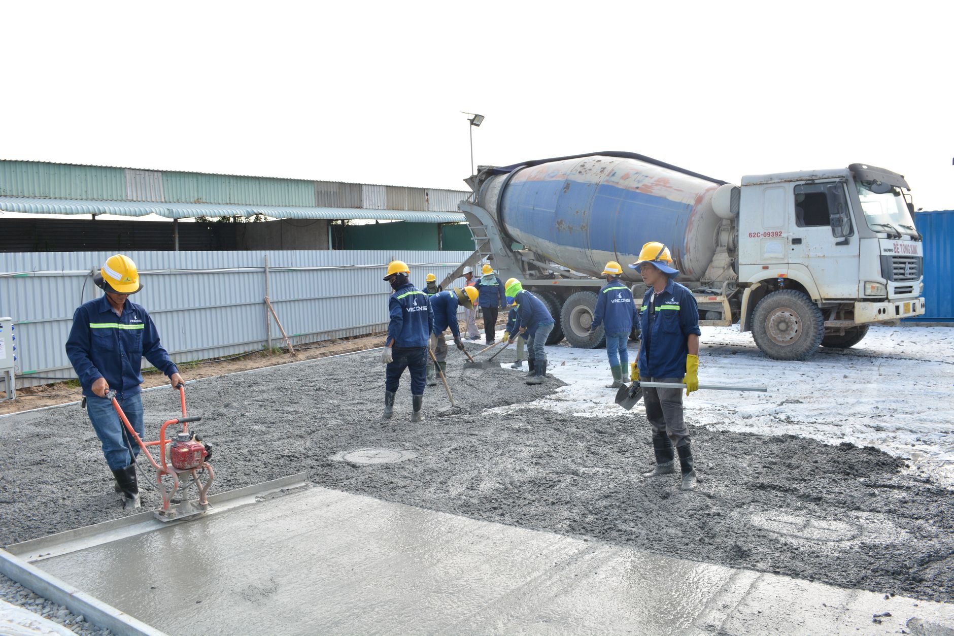 Ouvriers casqués lissant une dalle de béton frais avec une règle vibrante, devant un camion toupie sur un chantier.