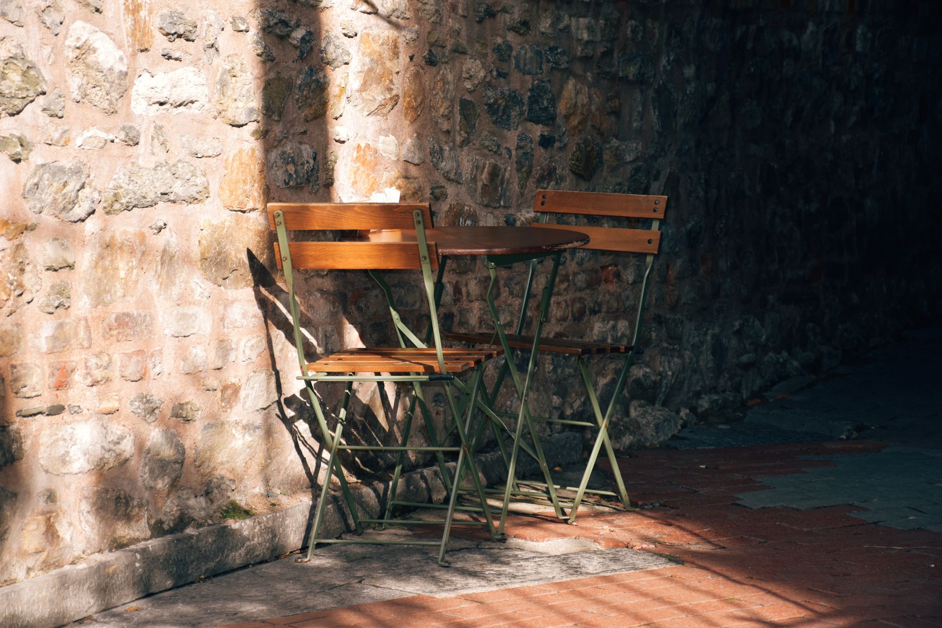 Table ronde et deux chaises pliantes en bois et métal vert, installées contre un mur de pierres, dans une lumière contrastée.